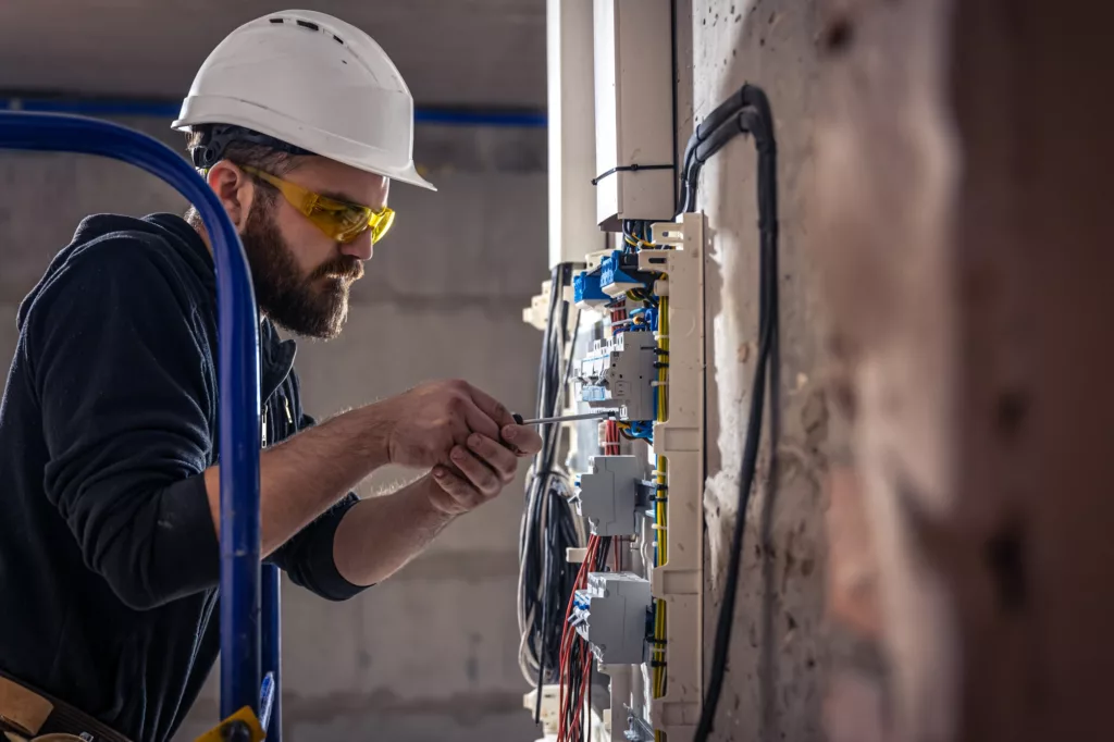 A male electrician works in a switchboard with an electrical connecting cable.